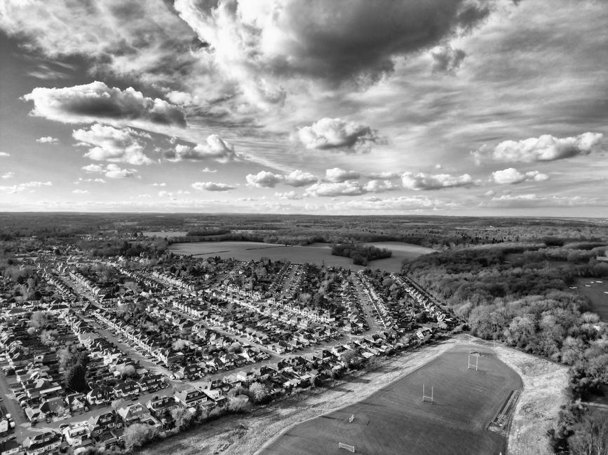 A black-and-white aerial photograph showcases a residential area with densely packed rows of houses and streets extending into the distance. In the foreground, there is a sports field bordered by a paved running track, with goalposts visible on the grassy area. Surrounding the residential neighborhood are patches of trees and open land, with a large body of water further back. The sky above is filled with scattered clouds, casting shadows across the landscape. This scene highlights the suburban environment and open spaces near Watford, compatible with house removals and moving logistics, illustrating the area's layout and environment as part of a relocation service overview by Watford Man and Van.