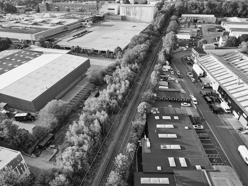 A black and white aerial photograph of a commercial or industrial area showing several large flat-roofed buildings with parking lots filled with vehicles, some delivery vans and cars. A dense line of trees runs parallel to railway tracks that pass through the middle of the scene, separating the parking areas from a warehouse or factory building. The image captures external spaces with vehicles being loaded or unloaded, and the surrounding environment includes additional warehouses and industrial units. The context suggests a busy environment where furniture and household items could be packaged and transported as part of home relocation or moving services, reflecting the logistics involved in packing and loading processes, as operated by companies like Watford Man and Van.