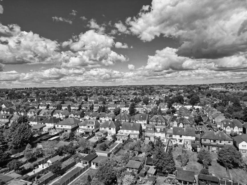 A black-and-white aerial photograph showcases a residential area with densely packed rows of houses and streets extending into the distance. In the foreground, there is a sports field bordered by a paved running track, with goalposts visible on the grassy area. Surrounding the residential neighborhood are patches of trees and open land, with a large body of water further back. The sky above is filled with scattered clouds, casting shadows across the landscape. This scene highlights the suburban environment and open spaces near Watford, compatible with house removals and moving logistics, illustrating the area's layout and environment as part of a relocation service overview by Watford Man and Van.